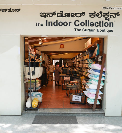 The storefront of 'The Indoor Collection' curtain boutique, showing a wide opening with customers inside and pillows displayed on racks on the right side.